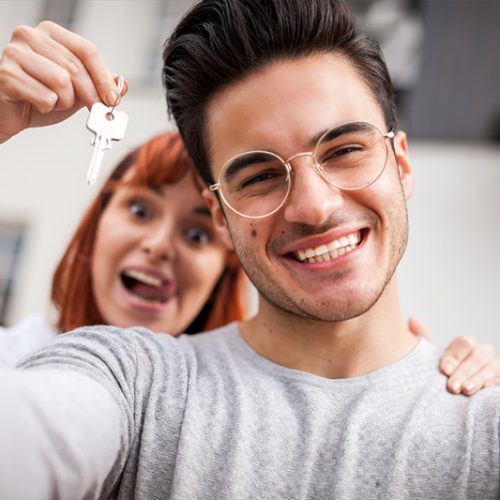Home 8 A smiling young man takes a selfie while a woman behind him excitedly holds up keys, suggesting they have just acquired a new home. Both appear happy and celebratory.