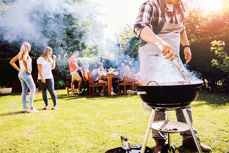 Home 2 A person grilling food on a barbecue in a sunny backyard while a group of people socialize and sit at a table in the background. Smoke rises from the grill, and trees and grass surround the scene.