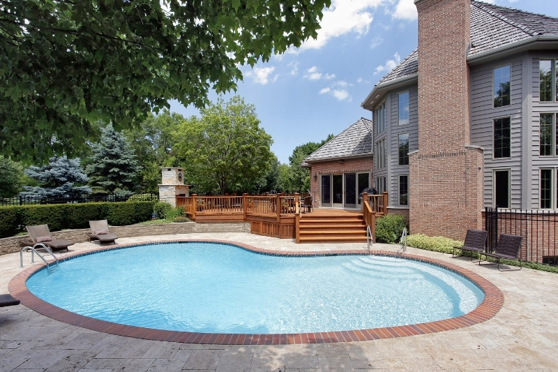 A kidney-shaped swimming pool with a brick border in a backyard, surrounded by a patio, wooden deck, lounge chairs, and a large brick house with tall windows and a chimney. Lush greenery and trees are in the background.