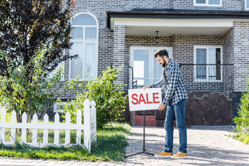 A man places a SALE sign in front of a modern brick house with a white fence and green lawn.