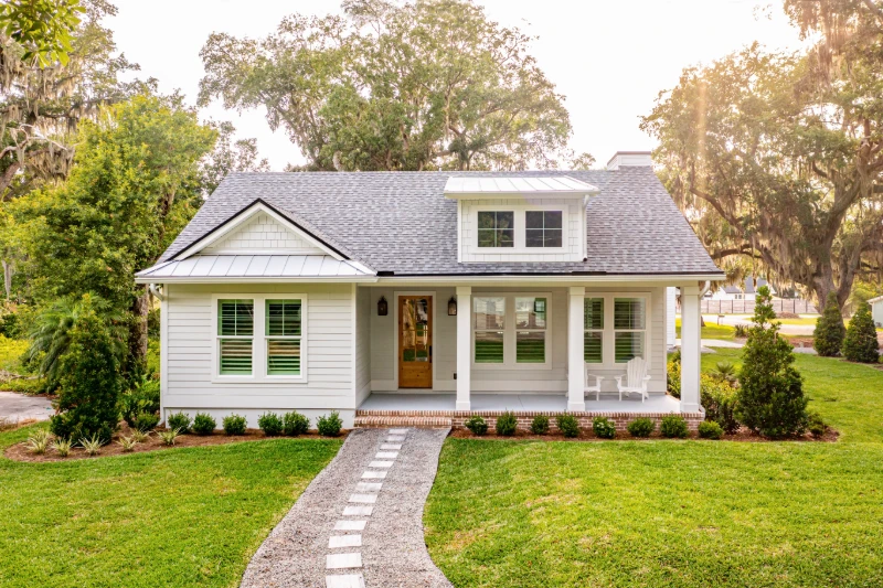 A charming white cottage with a front porch, brick steps, and two rocking chairs, surrounded by green grass, shrubs, and tall trees on a sunny day.